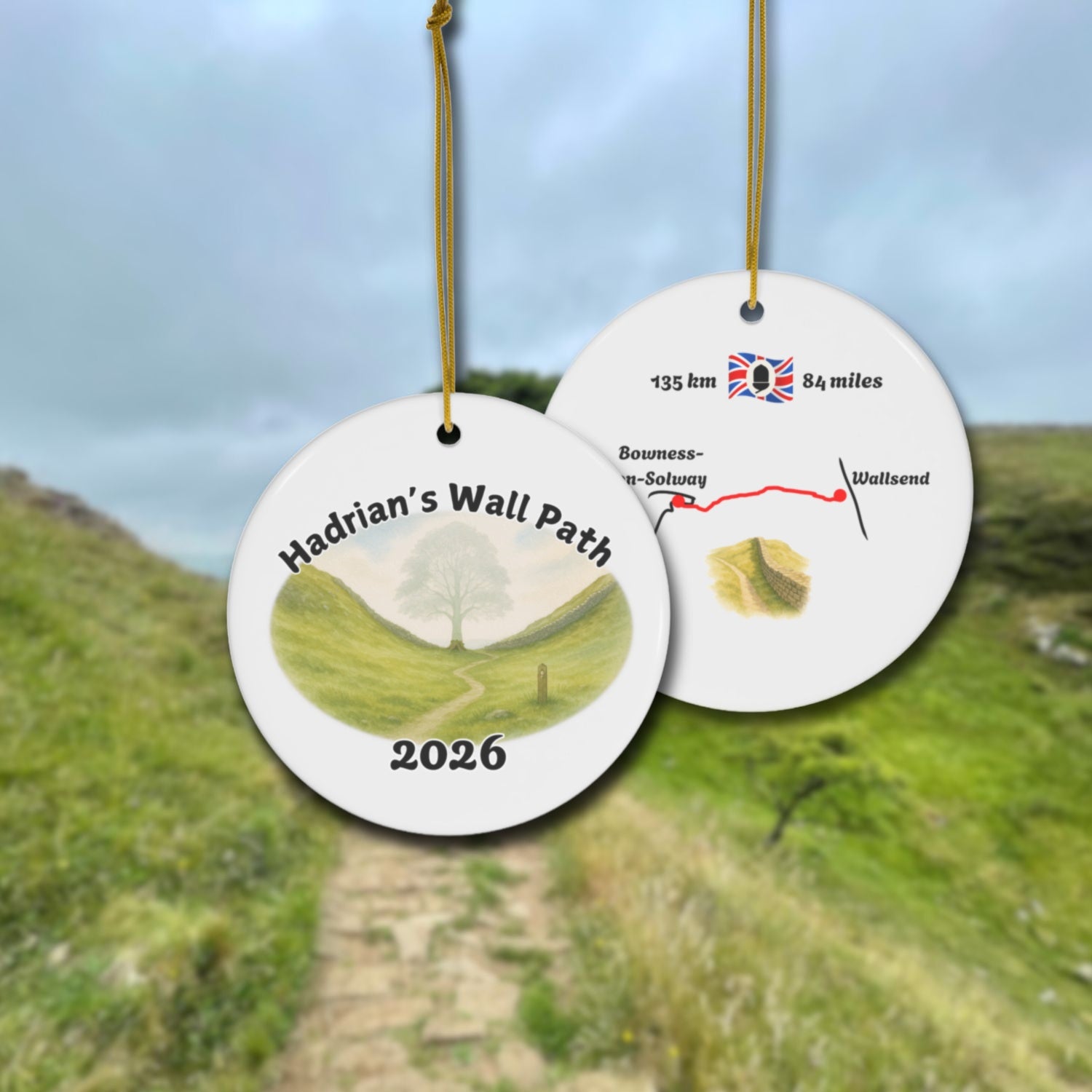 Hadrian's Wall Path ceramic ornament with watercolor landscape shown front and back in front of trail landscape.