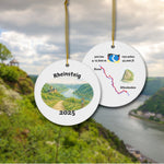 Front and back of the Rheinsteig keepsake ornament with vineyard watercolor scene and stylized trail map. Shown against a scenic Rhine Valley backdrop.
